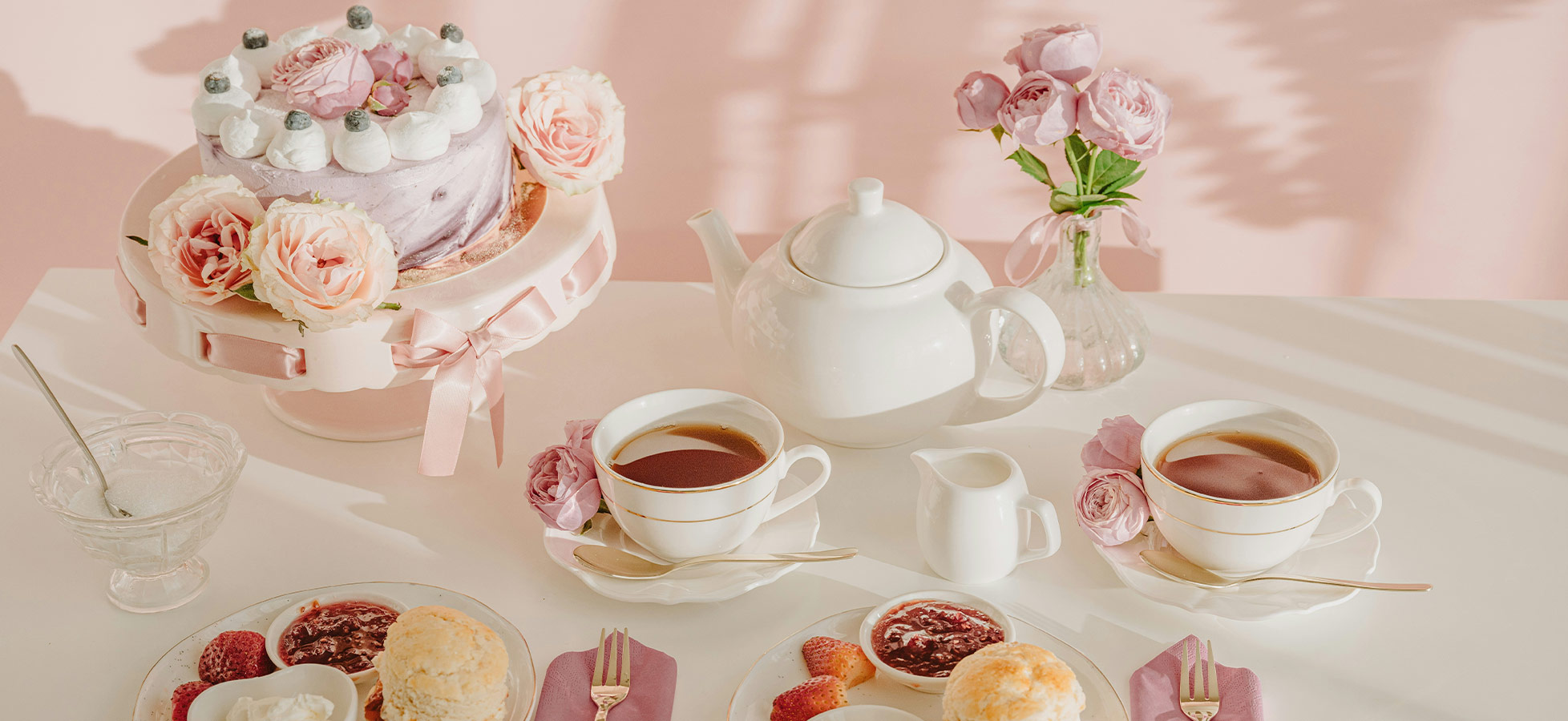 Auf dem Tisch sind zwei Tassen Tee, eine Teekanne und eine kleine Milchkanne platziert. Neben den Teetassen liegen Teller mit Gebäck, Marmelade und frischen Erdbeeren. Im Hintergrund steht eine dekorierte Torte mit Rosen, und eine Vase mit Rosenblüten ergänzt die fröhliche Atmosphäre.