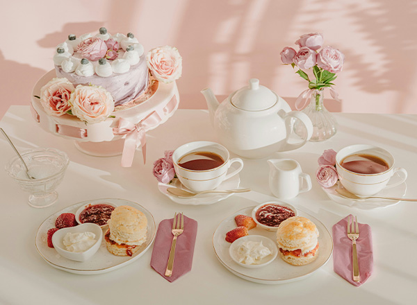 Tischgedeck mit einem großen, mehrstöckigen Kuchen, umgeben von rosa Rosen. Auf dem Tisch stehen zwei Porzellantassen mit Tee, ein Teekännchen, Sahne und zwei Teller mit Scones, einer mit Erdbeermarmelade, der andere mit Sahne.