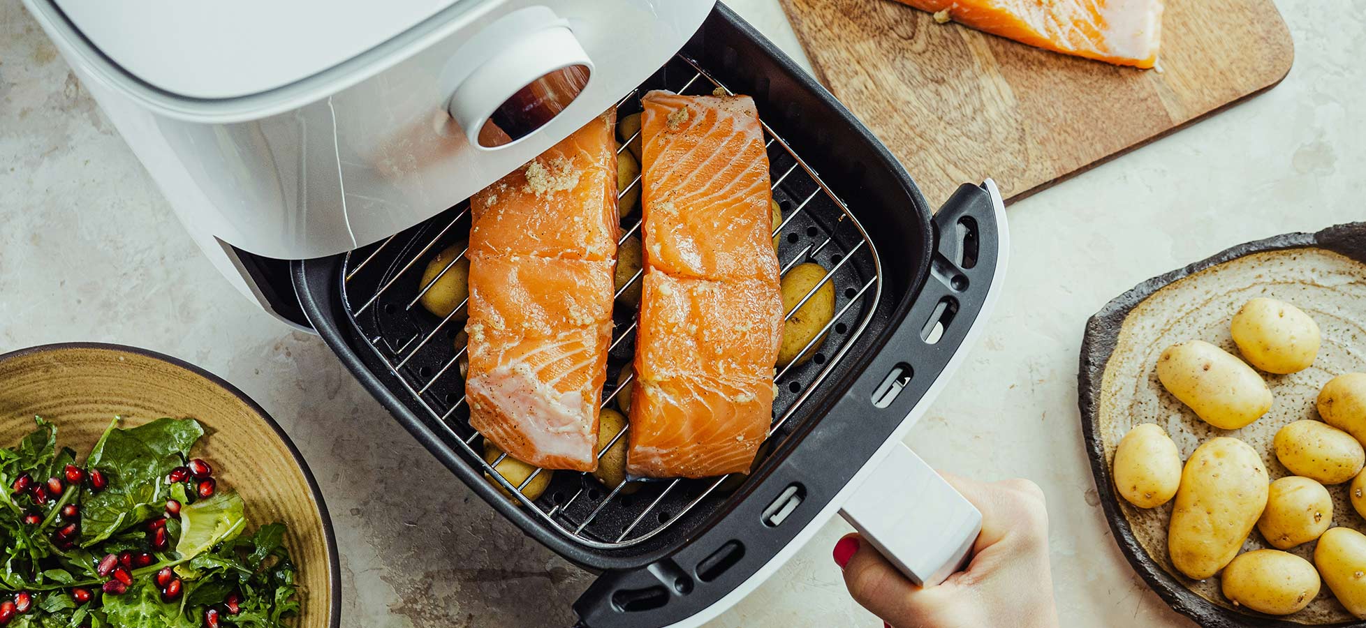 Ein Luftfritteuse mit zwei Lachsfilets auf einem Grillrost. Neben der Fritteuse liegen rohe Kartoffeln auf einem Teller und frischer Salat in einer Schüssel. Die Hand einer Person greift nach dem Griff der Fritteuse.