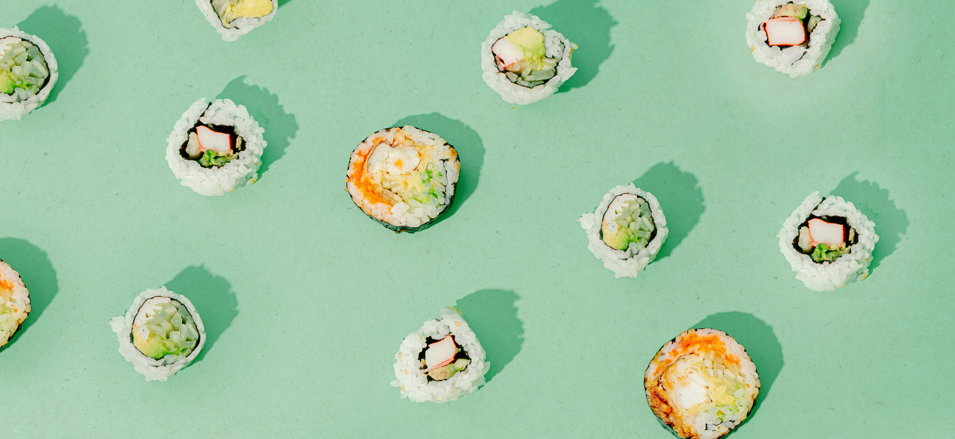 Ein flaches, mintgrünes Hintergrundbild zeigt verschiedene Sushi-Rollen. Die Rollen sind in unterschiedlichen Formen und Farben angeordnet, mit sichtbarem Reis, Algen und verschiedenen Füllungen wie Gemüse und Fisch.