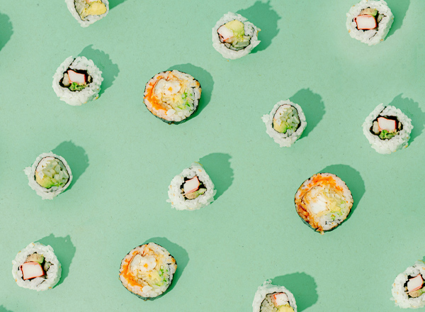 Auf einem mintgrünen Hintergrund sind verschiedene Sushi-Rollen in einer ruhigen Anordnung verteilt. Die Sushi-Rollen enthalten Zutaten wie Gemüse und Fisch, einige sind mit einer orangefarbenen oder weißen Füllung sichtbar. Das Bild vermittelt ein frisches und einladendes Gefühl.