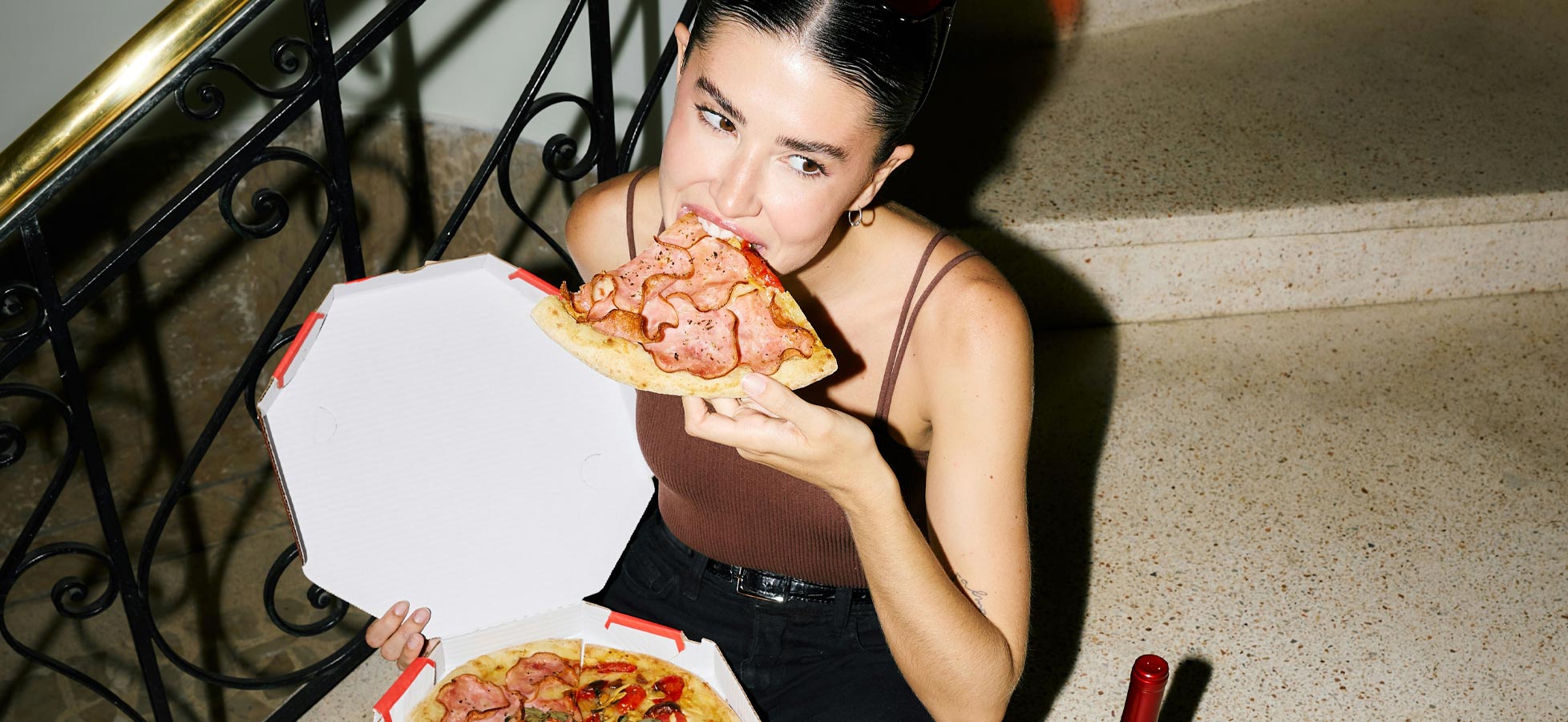 Eine Person sitzt auf einer Treppe und hält eine Pizzaschachtel offen. Sie beißt in ein Stück Pizza, die mit Schinken belegt ist. Die Atmosphäre wirkt entspannt und lässig.