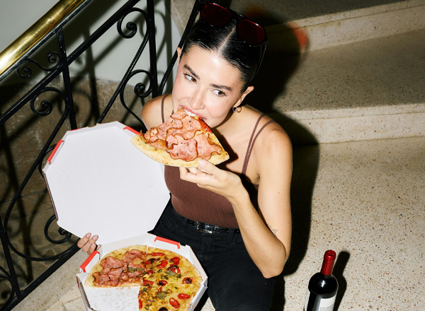 Eine junge Frau sitzt auf einer Treppe und beißt in ein Stück Pizza mit Schinken. Vor ihr steht eine Pizzaschachtel mit einer weiteren Pizza. Neben ihr steht eine Flasche Wein. Sie trägt eine schwarze Hose und ein braunes Oberteil.