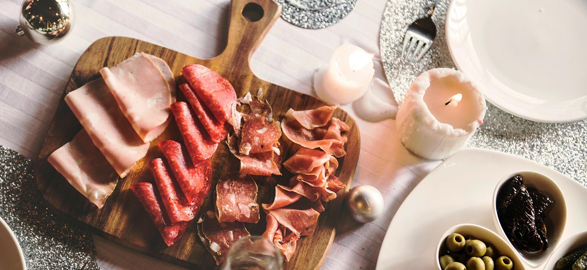 Ein Holzbrett ist mit verschiedenen Wurstsorten garniert, darunter Schinken und Salami. Daneben befinden sich eine Kerze und kleine Schüsseln mit Oliven. Der Tisch ist festlich gedeckt, was auf eine besondere Gelegenheit hinweist.