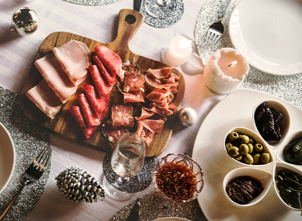 Die Abbildung zeigt eine festlich gedeckte Tafel mit einer Holzplatte, auf der verschiedene Wurst- und Schinkenarten präsentiert sind. Dazu stehen Gläser und kleine Schalen mit Oliven und anderen Snacks. Einige Kerzen sorgen für eine stimmungsvolle Beleuchtung.
