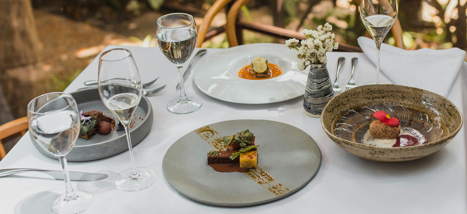 Ein eleganter Tisch ist gedeckt mit verschiedenen Tellergerichten, darunter Desserts und Hauptspeisen, umgeben von Gläsern mit Wasser und Sekt. In der Mitte steht eine kleine Vase mit frischen Blumen. Das Ambiente ist hell und freundlich, mit Pflanzen im Hintergrund.