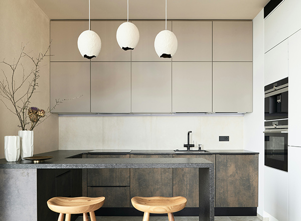 Eine moderne Küche mit einem schwarzen Küchenblock und hellen Wänden. Über der Arbeitsfläche hängen drei runde, weiße Lampen. Auf dem Tisch stehen zwei weiße Vasen mit Zweigen. Zwei Holzstühle sind vor dem Küchenblock platziert.