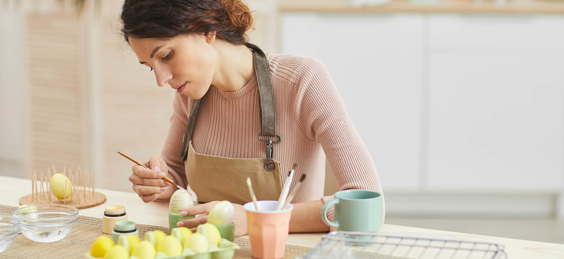 Eine Frau mit einem braunen Schurz sitzt an einem Tisch und bemalt ein Ei mit einem Pinsel. Vor ihr sind farbige Eier, ein Glas mit Wasser und verschiedene Malutensilien zu sehen. Der Hintergrund ist hell und zeigt eine moderne Küche.