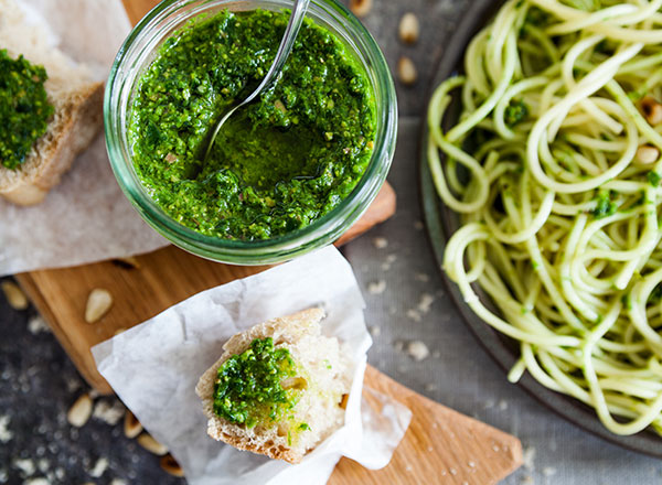 Ein Glas Pesto mit einem Löffel darin steht neben einer mit Pesto bestrichenen Brotscheibe, im Hintergrund ist ein Teller mit Pasta mit Pesto zu sehen.