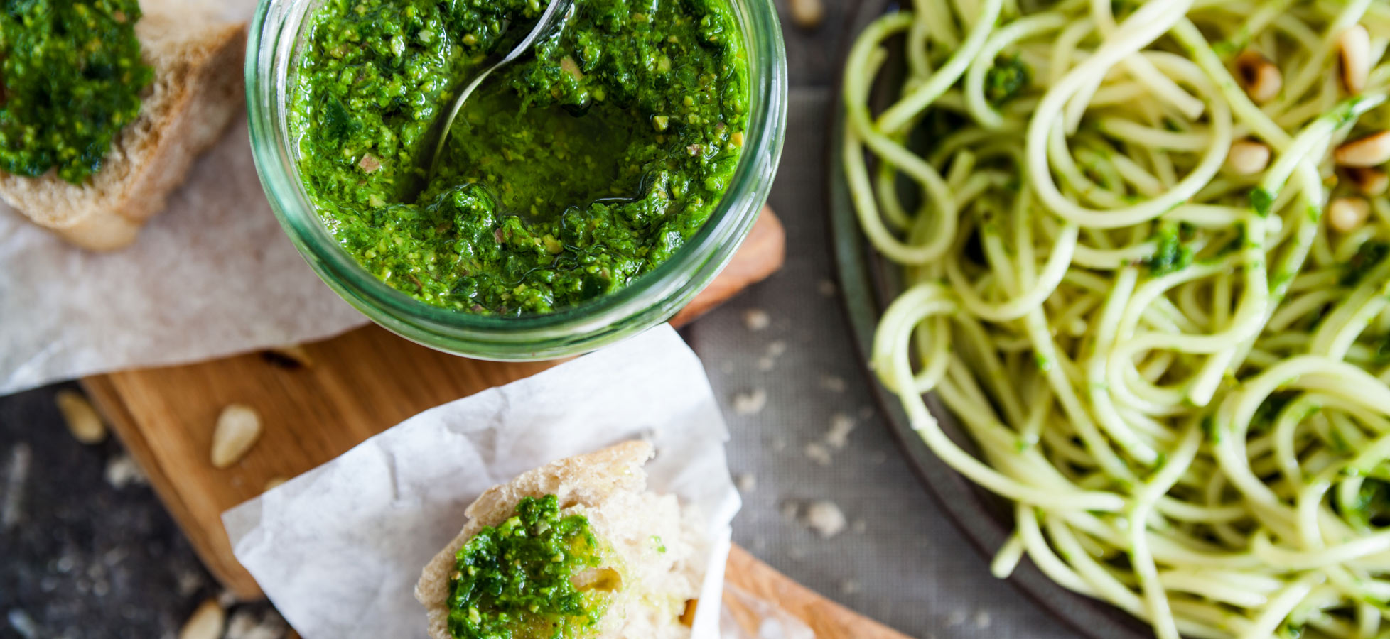 Ein Glas Pesto mit einem Löffel darin steht neben einer mit Pesto bestrichenen Brotscheibe, im Hintergrund ist ein Teller mit Pasta mit Pesto zu sehen.