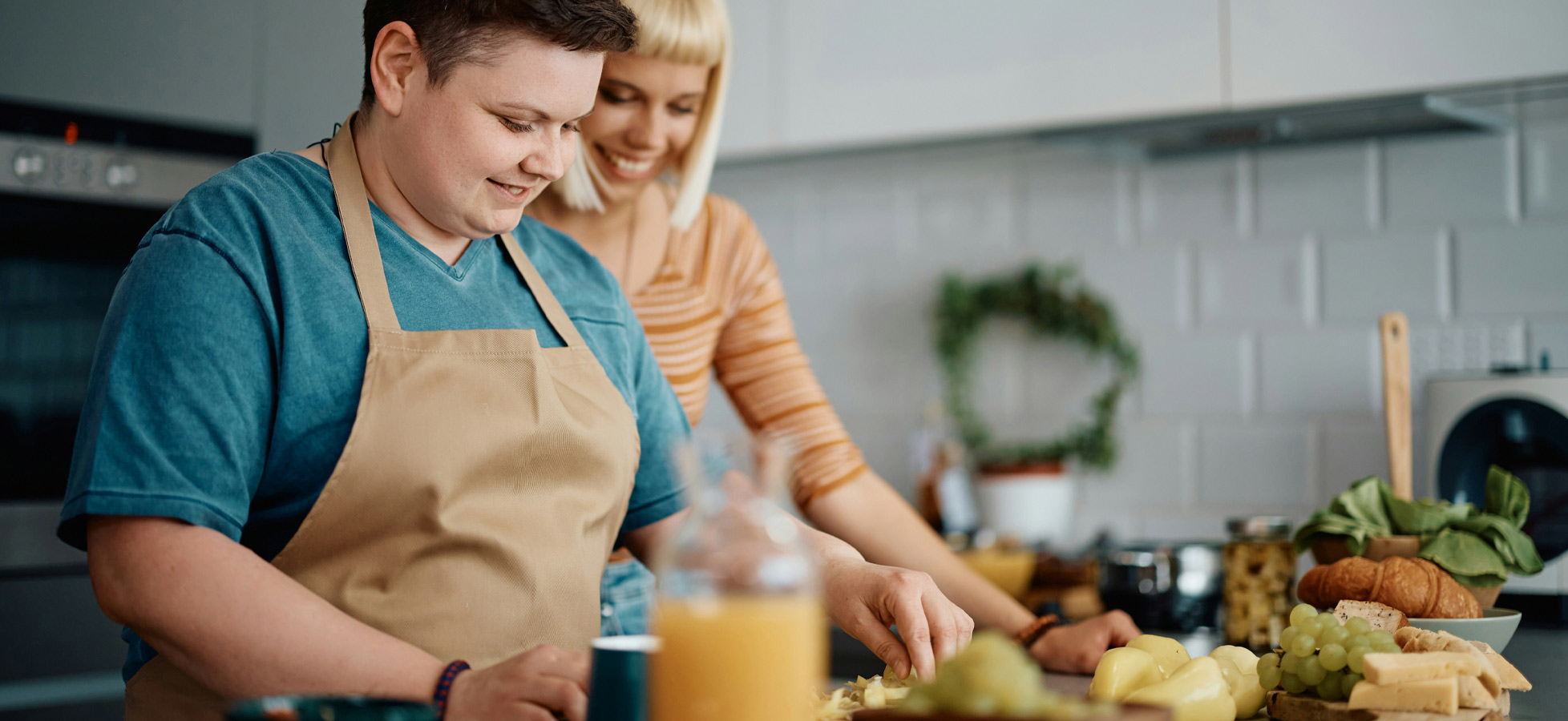 Zwei Personen in einer modernen Küche bereiten gemeinsam eine Mahlzeit vor. Eine Person trägt eine Schürze und schneidet Zutaten, während die andere lächelt und sie unterstützt. Auf der Arbeitsfläche sind verschiedene Lebensmittel sichtbar, darunter Obst und Gemüse.
