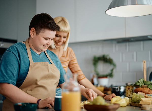 Zwei Personen in einer modernen Küche bereiten gemeinsam eine Mahlzeit vor. Eine Person trägt eine Schürze und schneidet Zutaten, während die andere lächelt und sie unterstützt. Auf der Arbeitsfläche sind verschiedene Lebensmittel sichtbar, darunter Obst und Gemüse.