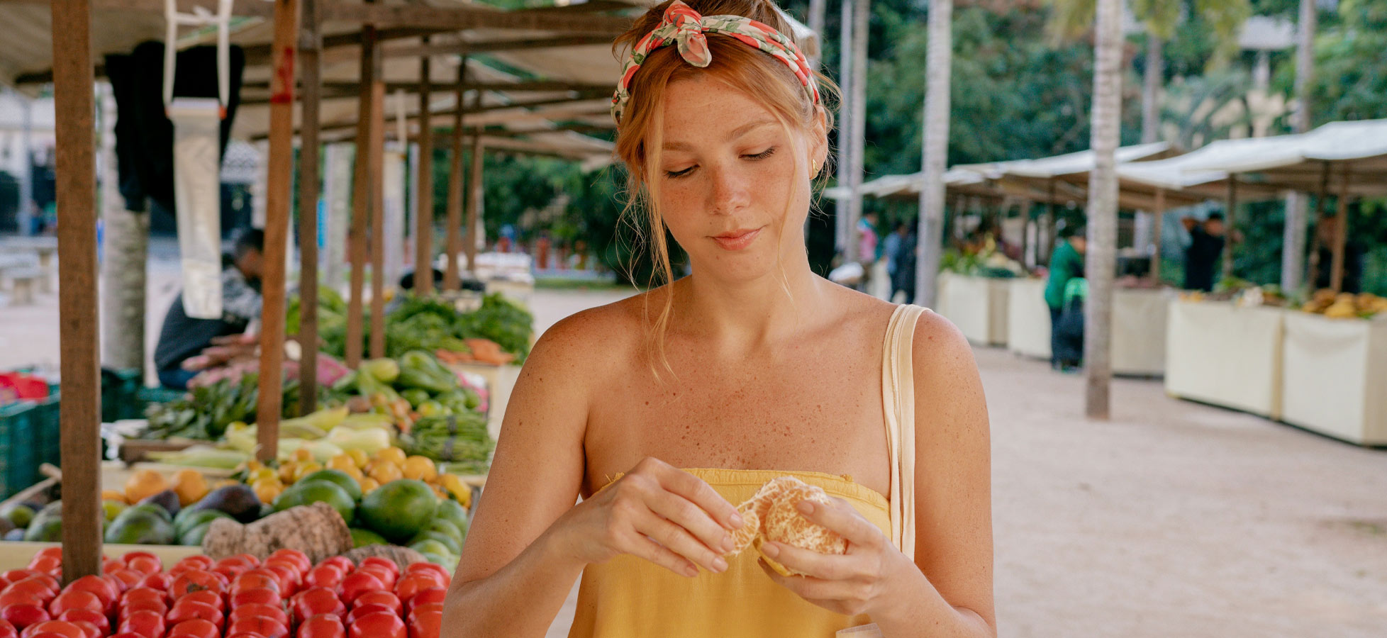 Eine Frau in einem gelben Top steht an einem Marktstand, umgeben von frischem Obst und Gemüse. Sie hält ein Stück Obst in der Hand und schaut konzentriert darauf. Im Hintergrund sind weitere Marktstände und Menschen zu sehen.