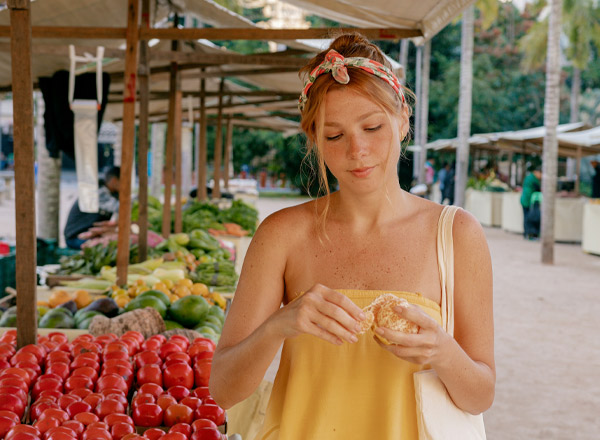 Eine Frau in einem gelben Top steht an einem Marktstand, umgeben von frischem Obst und Gemüse. Sie hält ein Stück Obst in der Hand und schaut konzentriert darauf. Im Hintergrund sind weitere Marktstände und Menschen zu sehen.