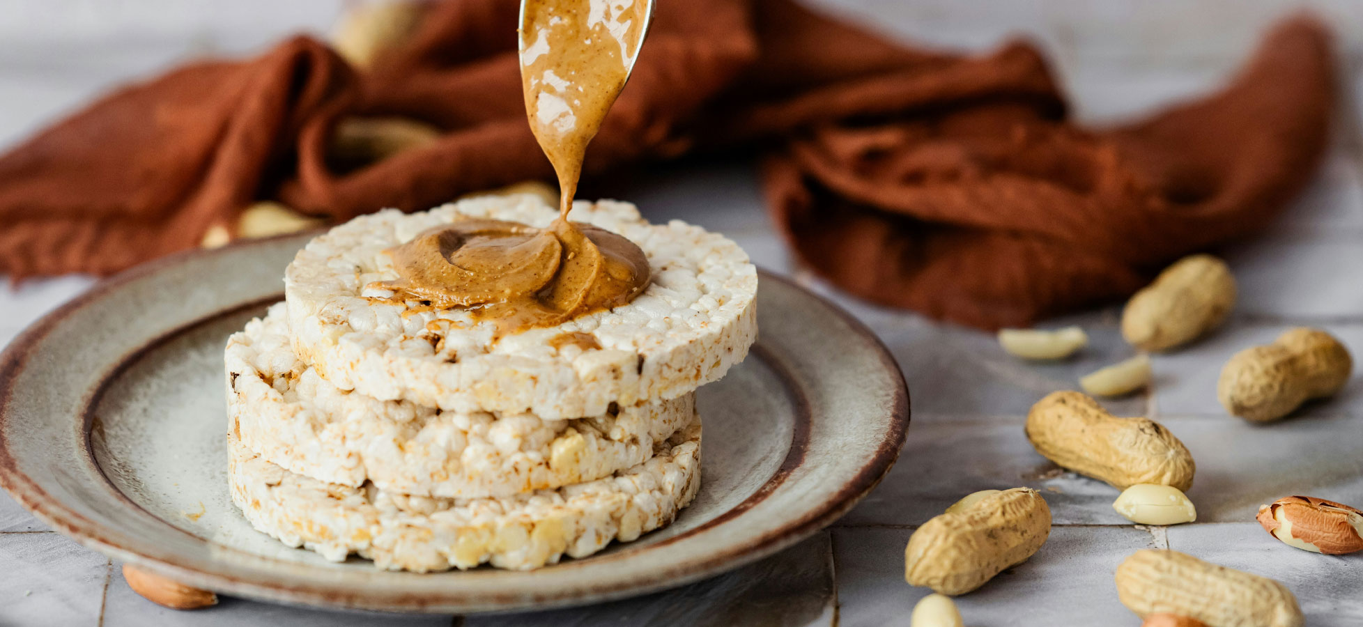 Auf einem Teller liegen mehrere Reiswaffeln, die mit Erdnussbutter bestrichen sind. Ein Löffel hebt die Erdnussbutter an, während im Hintergrund einige ungeschälte Erdnüsse sichtbar sind. Die Szene vermittelt einen gesunden Snack.