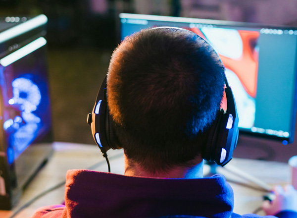 Ein Mensch sitzt mit einem Headset vor zwei Computermonitoren. Der Betrachter sieht lediglich den Hinterkopf und die Schultern des Nutzers. Die Monitore zeigen möglicherweise grafische Inhalte oder Anwendungen. Der Raum ist schwach beleuchtet, wodurch eine konzentrierte Atmosphäre entsteht.