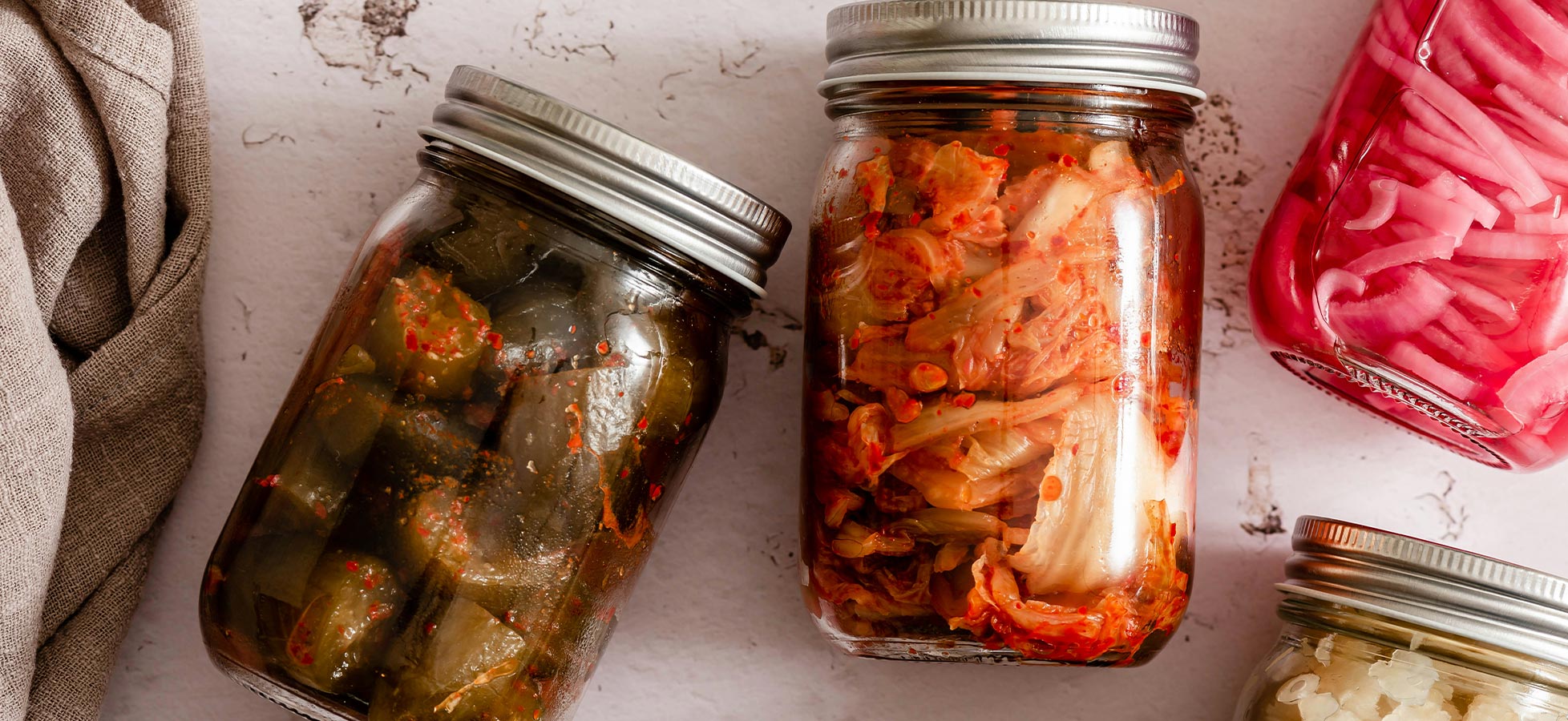 Drei Einmachgläser mit fermentierten Lebensmitteln auf einem Tisch. Links ein Glas mit grünen, scharfen eingelegten Gemüsesorten. Rechts ein Glas mit kimchi, das aus gehacktem Gemüse und Gewürzen besteht. Im Hintergrund sind weitere Gläser mit eingelegten Lebensmitteln zu sehen.
