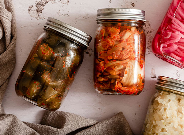 Drei Einmachgläser mit fermentierten Lebensmitteln auf einem Tisch. Links ein Glas mit grünen, scharfen eingelegten Gemüsesorten. Rechts ein Glas mit kimchi, das aus gehacktem Gemüse und Gewürzen besteht. Im Hintergrund sind weitere Gläser mit eingelegten Lebensmitteln zu sehen.