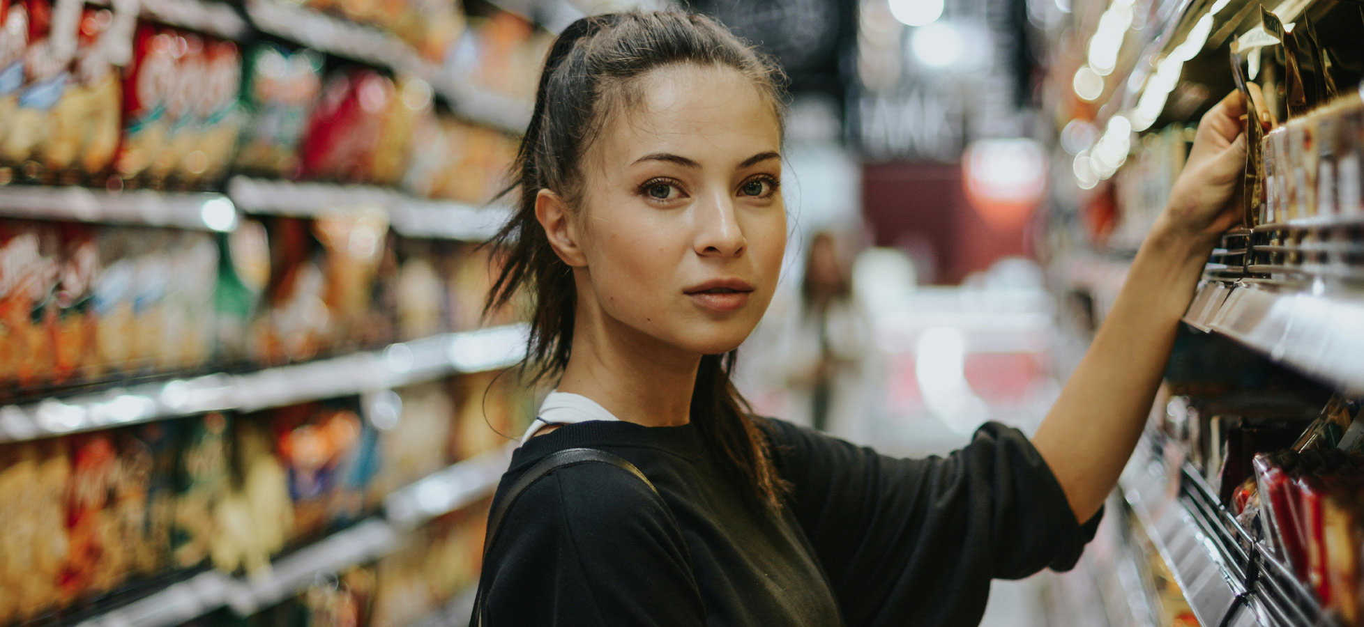 Eine junge Frau steht in einem Supermarktgang und betrachtet die Regale mit Lebensmitteln. Sie hat lange, dunkle Haare und trägt ein schwarzes Sweatshirt. Der Hintergrund zeigt eine Vielzahl von Verpackungen auf den Regalen, während andere Kunden im Hintergrund zu sehen sind.