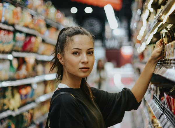 Eine junge Frau steht in einem Supermarktgang und betrachtet die Regale mit Lebensmitteln. Sie hat lange, dunkle Haare und trägt ein schwarzes Sweatshirt. Der Hintergrund zeigt eine Vielzahl von Verpackungen auf den Regalen, während andere Kunden im Hintergrund zu sehen sind.