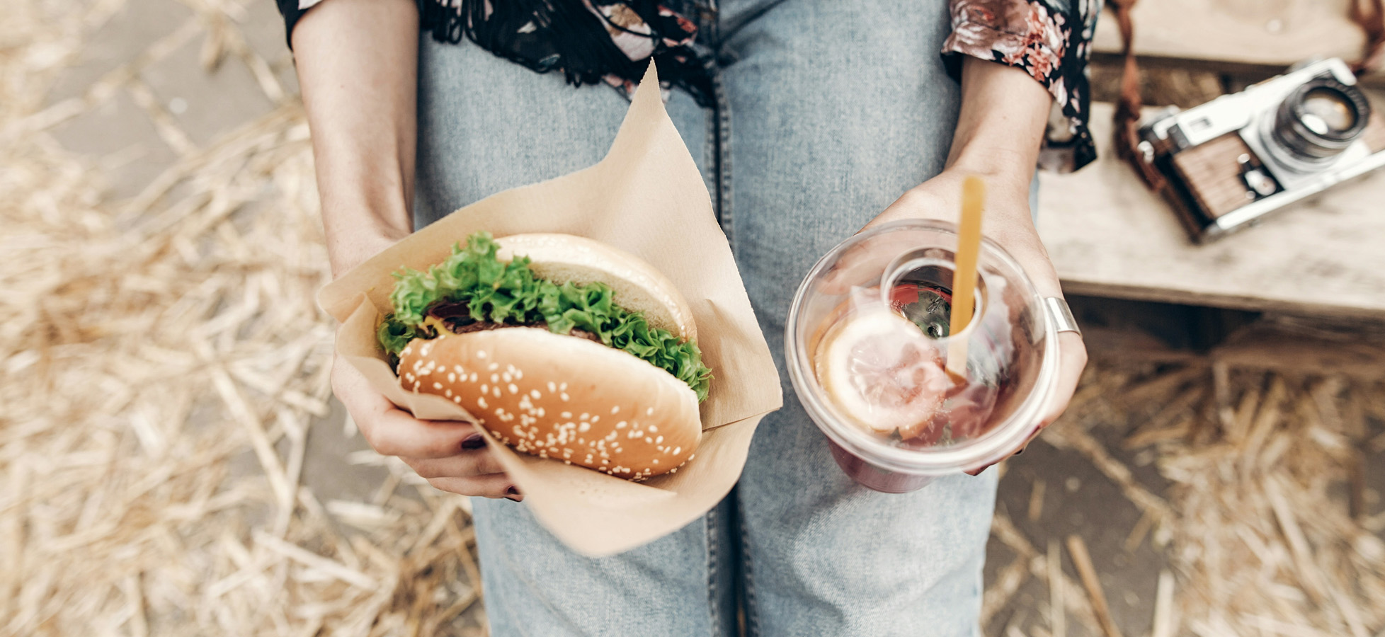 Eine Person hält einen Burger mit Sesambrötchen und frischem Salat in einer Papierunterlage in der einen Hand und ein Glas mit einem bunten Getränk und einer Zitronenscheibe in der anderen Hand. Der Hintergrund ist unscharf und enthält Stroh.