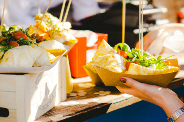 Eine Hand hält ein Pappschälchen mit gefülltem Teig und frischem Salat. Im Hintergrund stehen weitere Schalen mit ähnlichem Essen auf einem Tisch. Die Szenerie vermittelt eine einladende Atmosphäre eines Food-Markts.