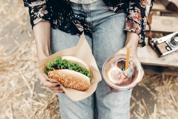 Eine Person sitzt auf einer Holzkiste und hält einen Hamburger mit Salat und ein Getränk in einem Becher mit Strohhalm in den Händen. Im Hintergrund ist eine verschwommene Kamera zu sehen.