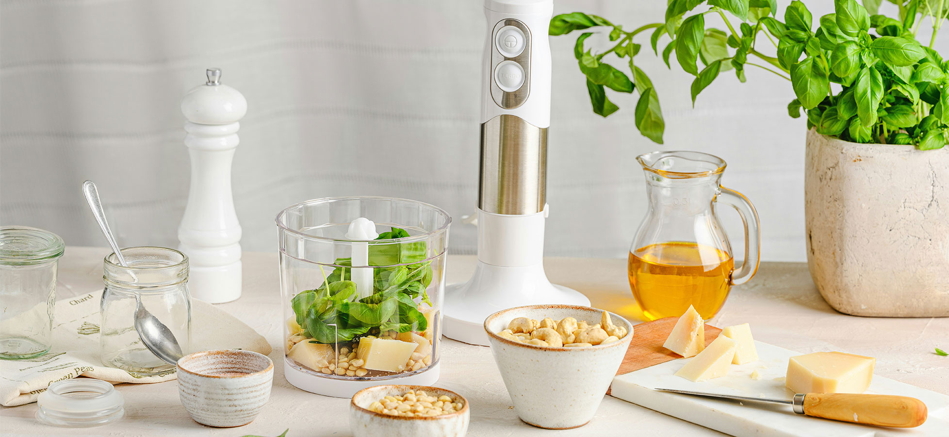 Eine Küchenanordnung mit einem weißen Handmixer, frischem Spinat, Butterstücken und Nüssen in Schalen. Daneben stehen ein Glas mit Öl und frisches Basilikum in einem Topf. Eine Salzstreuer und ein Schneidebrett mit Käse sind ebenfalls sichtbar.