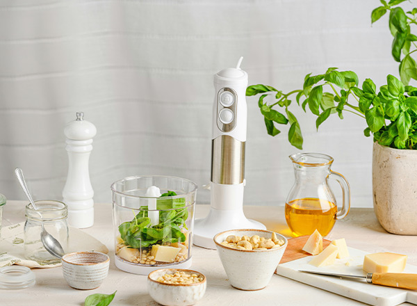 Eine Küchenanordnung mit einem weißen Handmixer, frischem Spinat, Butterstücken und Nüssen in Schalen. Daneben stehen ein Glas mit Öl und frisches Basilikum in einem Topf. Eine Salzstreuer und ein Schneidebrett mit Käse sind ebenfalls sichtbar.