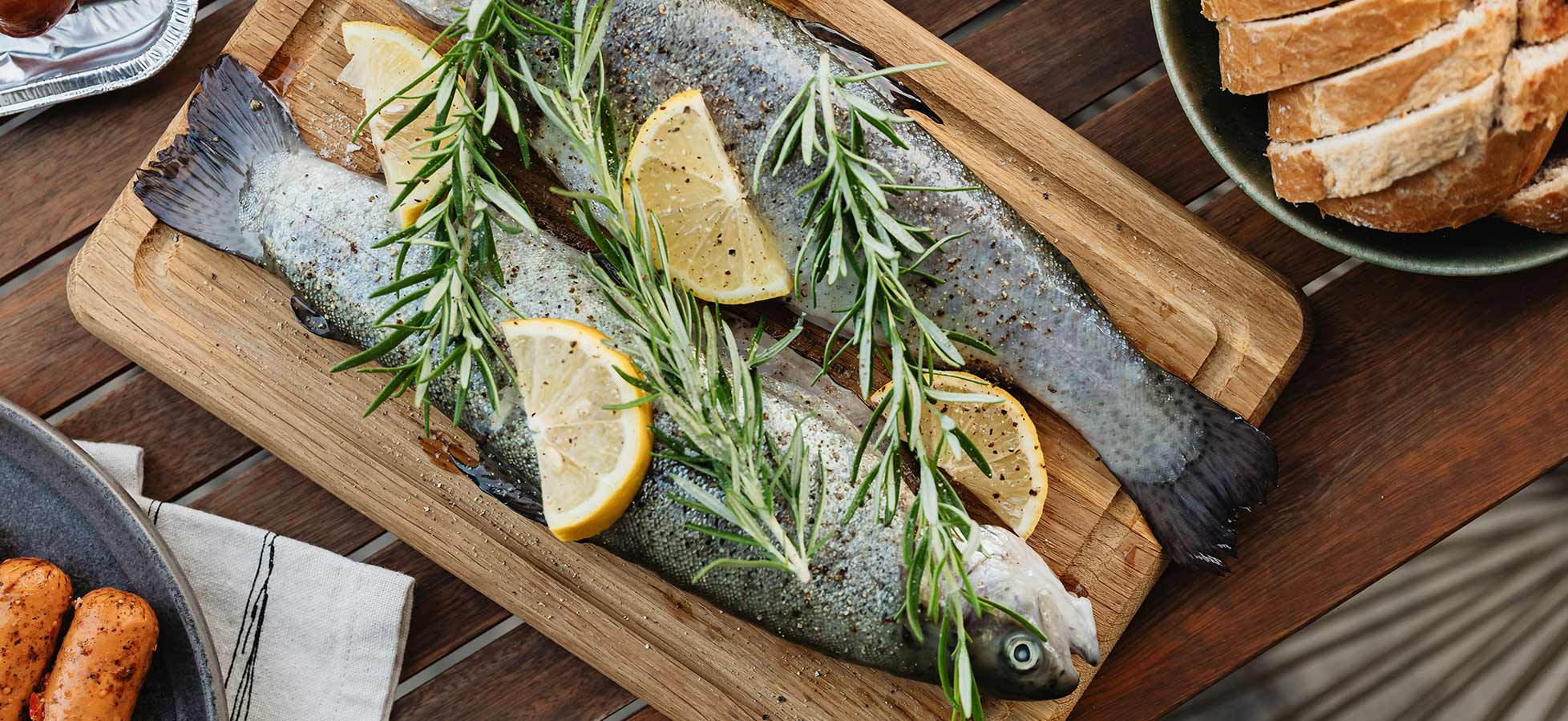Auf einem Holzbrett liegen zwei frische Fische, garniert mit Zitronenscheiben und Kräutern. Im Hintergrund sind einige Brotscheiben sichtbar. Die Anordnung vermittelt eine einladende, rustikale Atmosphäre für eine Mahlzeit.