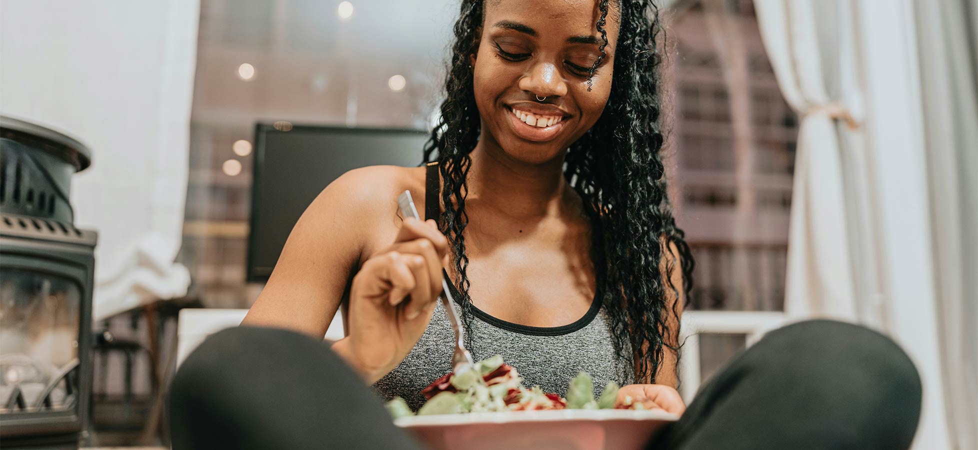 Eine lächelnde Frau sitzt auf dem Boden und genießt einen großen Salat aus einer Schüssel. Sie trägt ein sportliches Oberteil und hat lange, gewellte Haare. Der Raum wirkt gemütlich und legt den Fokus auf ihr glückliches Essen.