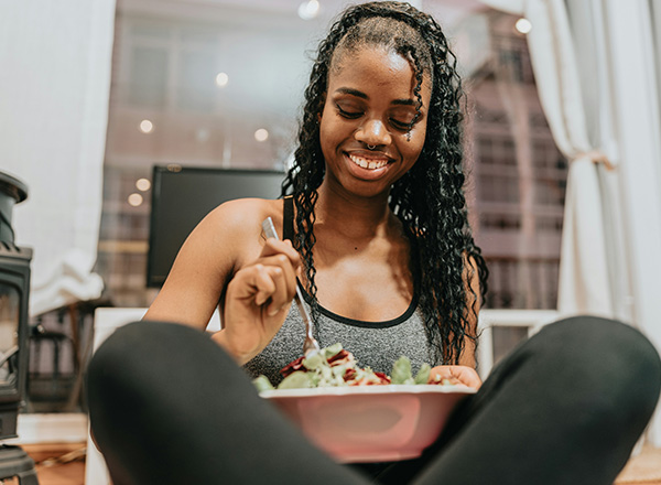 Eine junge Frau sitzt auf dem Boden und isst einen Salat aus einer großen Schüssel. Sie lächelt zufrieden, während sie mit einer Gabel einige Salatblätter aufnimmt. Im Hintergrund sind unscharfe Wohnraumelemente sichtbar.