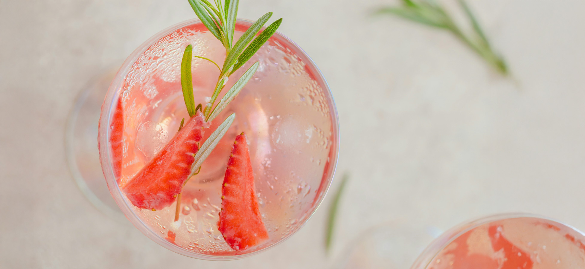 Ein Überblick über zwei Gläser Getränk mit Sprudel, in denen frische Erdbeerscheiben und ein Zweig Rosmarin schwimmen. Die Gläser stehen auf einer hellen, neutralen Oberfläche. Die Kombination vermittelt einen erfrischenden und aromatischen Eindruck.