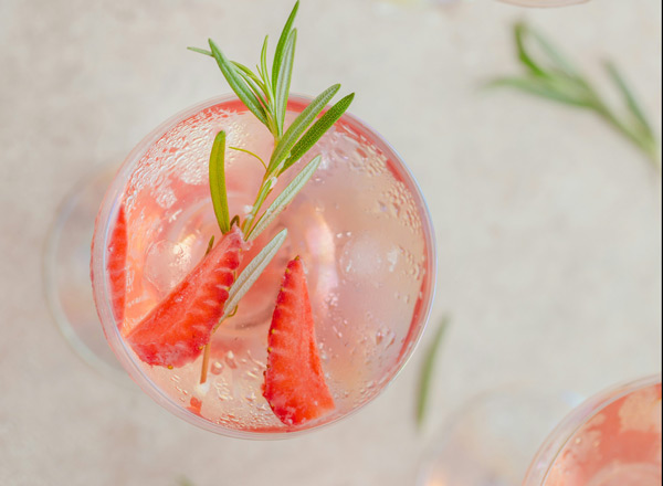 Ein Glas mit einem erfrischenden Getränk, in dem sich kleine Erdbeerscheiben und grüner Rosmarin befinden. Die Bläschen im Getränk lassen auf Kohlensäure schließen. Der Hintergrund ist hell und unscharf, was den Fokus auf das Glas lenkt.