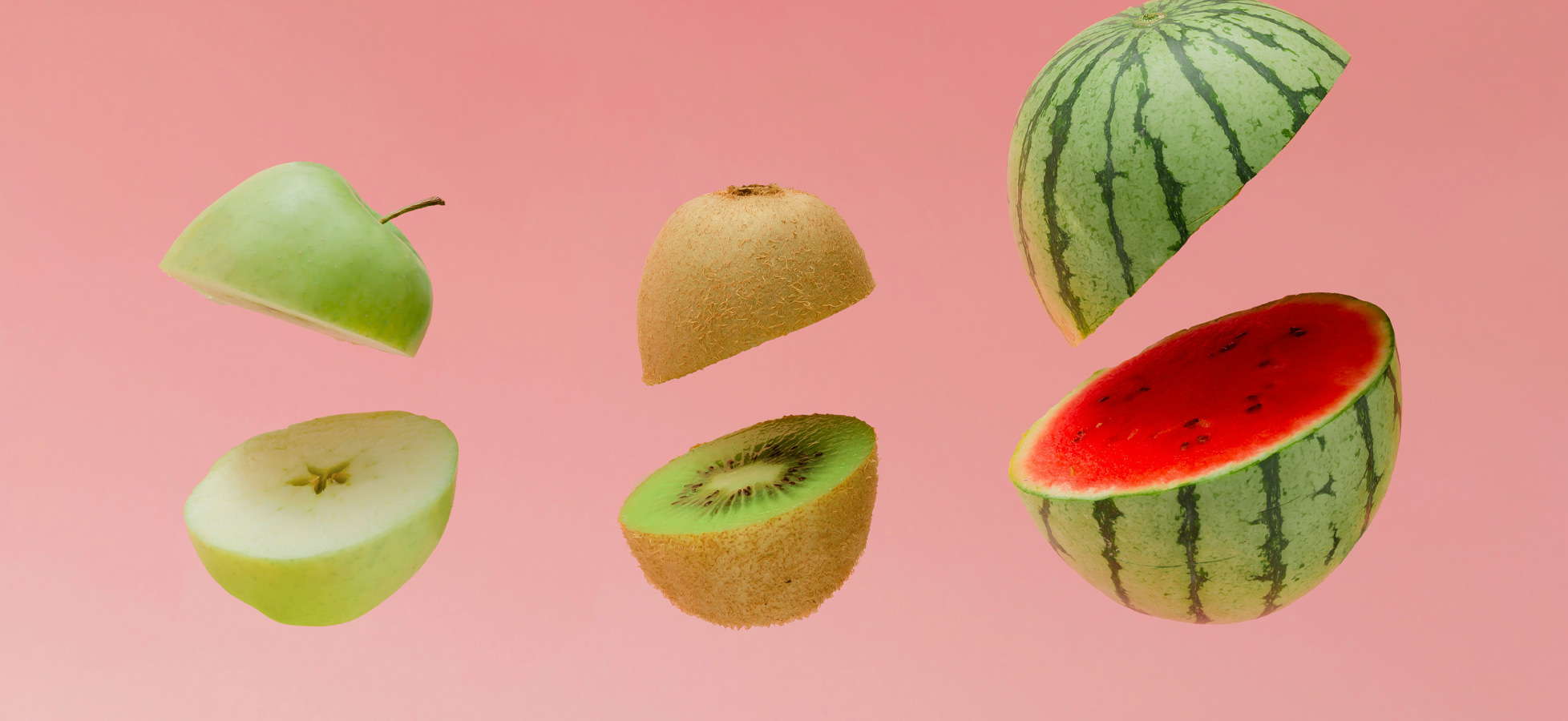 Eine Reihe von frischen Früchten schwebt vor einem rosa Hintergrund. Zu sehen sind ein grüner Apfel, ein halbierter Kiwi, eine ganze Wassermelone und ein halber Kiwi. Die Vielfalt der Früchte vermittelt ein frisches und einladendes Gefühl.