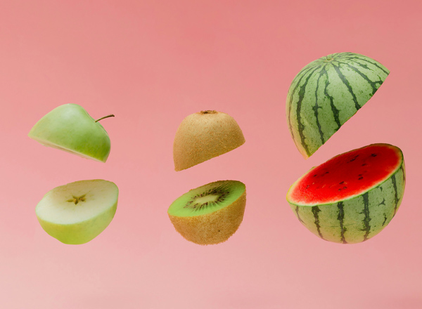 Eine Reihe von frischen Früchten schwebt vor einem rosa Hintergrund. Zu sehen sind ein grüner Apfel, ein halbierter Kiwi, eine ganze Wassermelone und ein halber Kiwi. Die Vielfalt der Früchte vermittelt ein frisches und einladendes Gefühl.