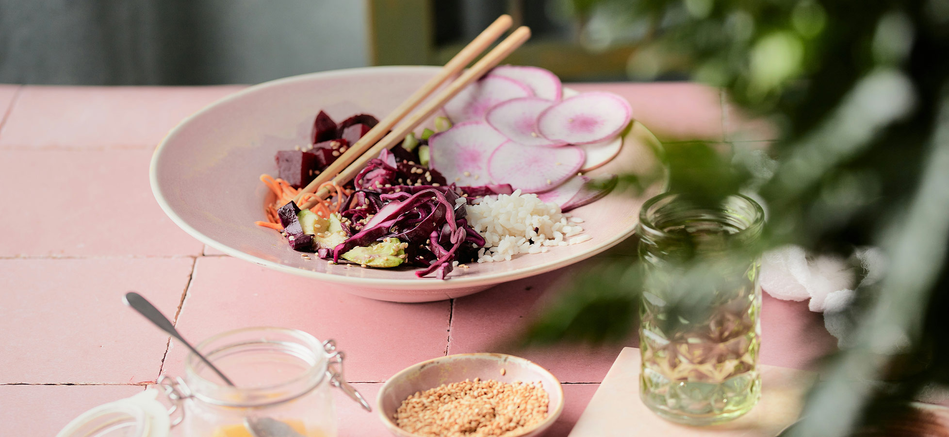Eine Schüssel mit buntem Salat, darunter Gemüse wie rote Bete, Karotten und Rotkohl, ergänzt durch Reis und Radieschenscheiben. Zwei Essstäbchen liegen auf dem Rand der Schüssel. Im Hintergrund ist ein Glas mit Getränk und kleine Behälter mit Zutaten sichtbar.