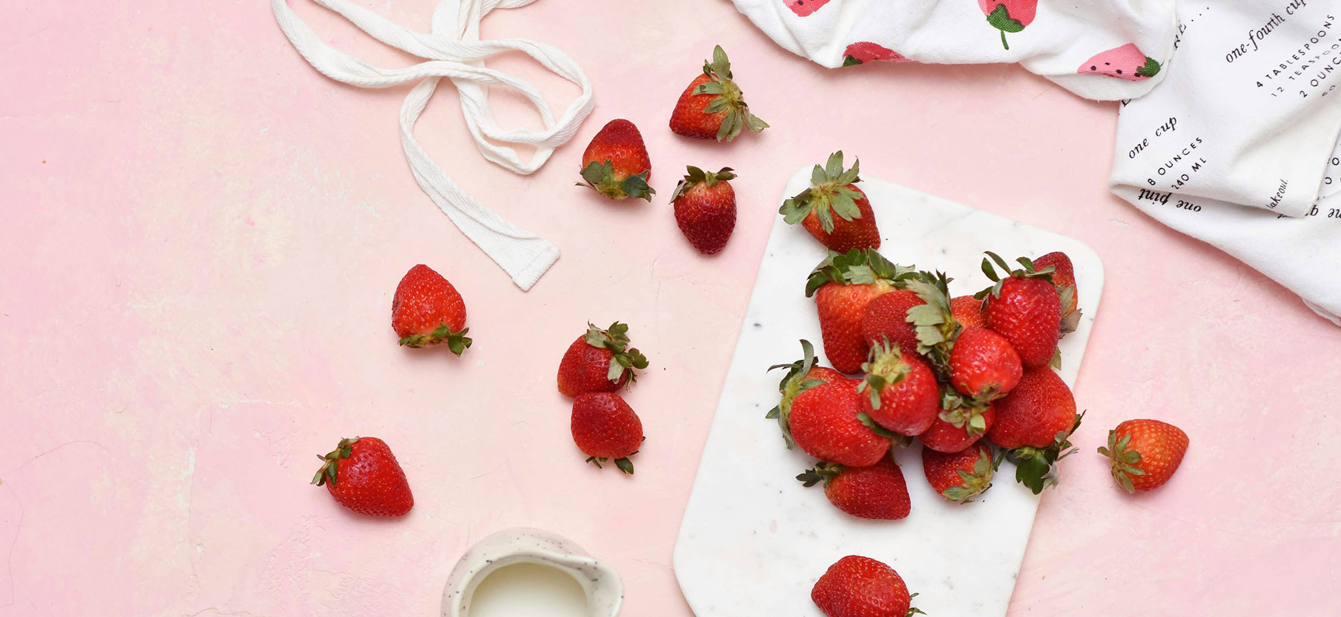 Frische Erdbeeren liegen auf einem weißen Schneidebrett. Einige Erdbeeren sind verstreut, und ein Becher mit Milch ist ebenfalls sichtbar. Der Hintergrund ist rosa, was eine fröhliche, einladende Atmosphäre schafft.
