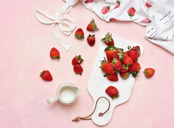 Ein Holzbrett ist mit frischen Erdbeeren bedeckt, einige liegen verstreut auf einer rosa Unterlage. Daneben steht eine kleine Kanne mit Milch. Im Hintergrund ist ein Handtuch mit Erdbeermuster zu sehen.