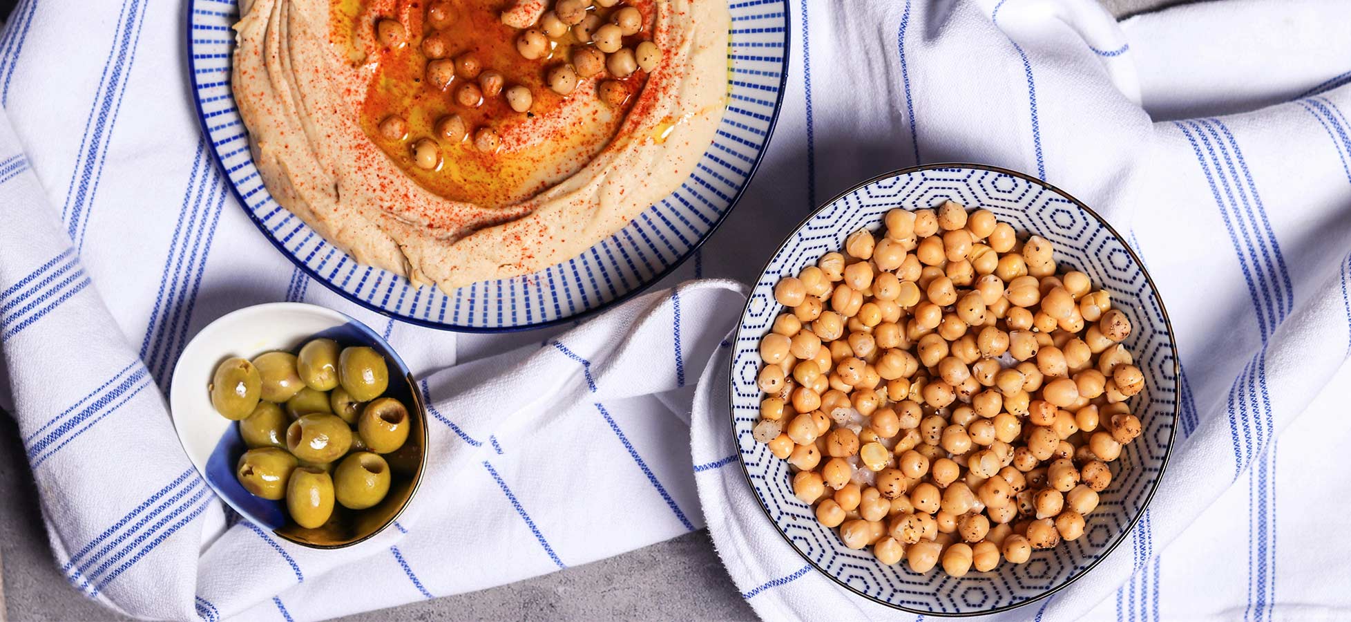 Ein Teller mit Hummus, garniert mit Kichererbsen und Paprikagewürz, steht auf einem blauen und weißen gestreiften Geschirrtuch. Neben dem Hummus befindet sich eine Schüssel mit ungewürzten Kichererbsen und eine kleine Schüssel mit grünen Oliven.