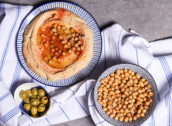 Ein Teller mit Hummus, garniert mit Kichererbsen und Paprika, steht auf einem Tisch. Daneben befinden sich zwei Schalen: eine mit grünen Oliven und eine mit ungeschälten Kichererbsen. Das Ganze liegt auf einem karierten Geschirrtuch.