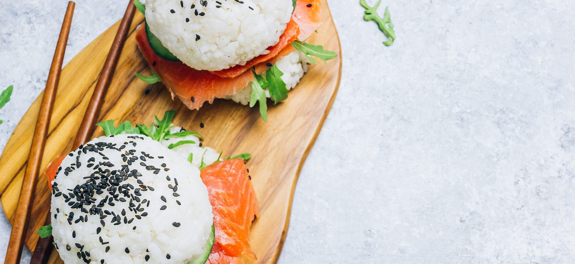 Zwei Sushi-ähnliche Snacks auf einem Holzbrett. Die Snacks bestehen aus Reis mit schwarzem Sesam, gefüllt mit Räucherlachs und Rucola. Neben den Snacks liegen Essstäbchen. Der Hintergrund ist hell und unauffällig.