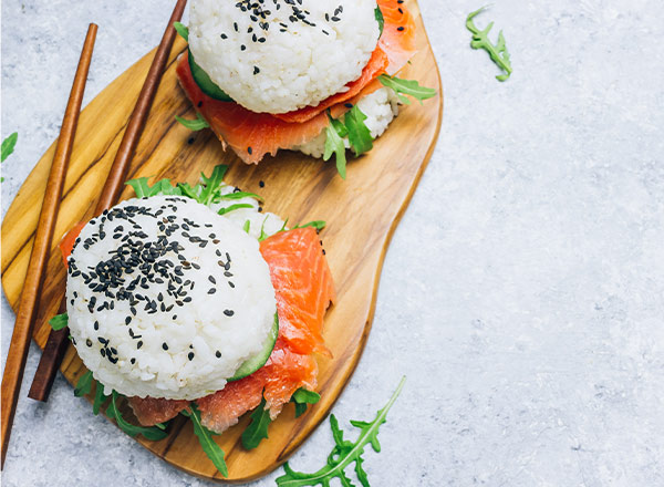 Zwei Sushi-ähnliche Snacks auf einem Holzbrett. Die Snacks bestehen aus Reis mit schwarzem Sesam, gefüllt mit Räucherlachs und Rucola. Neben den Snacks liegen Essstäbchen. Der Hintergrund ist hell und unauffällig.