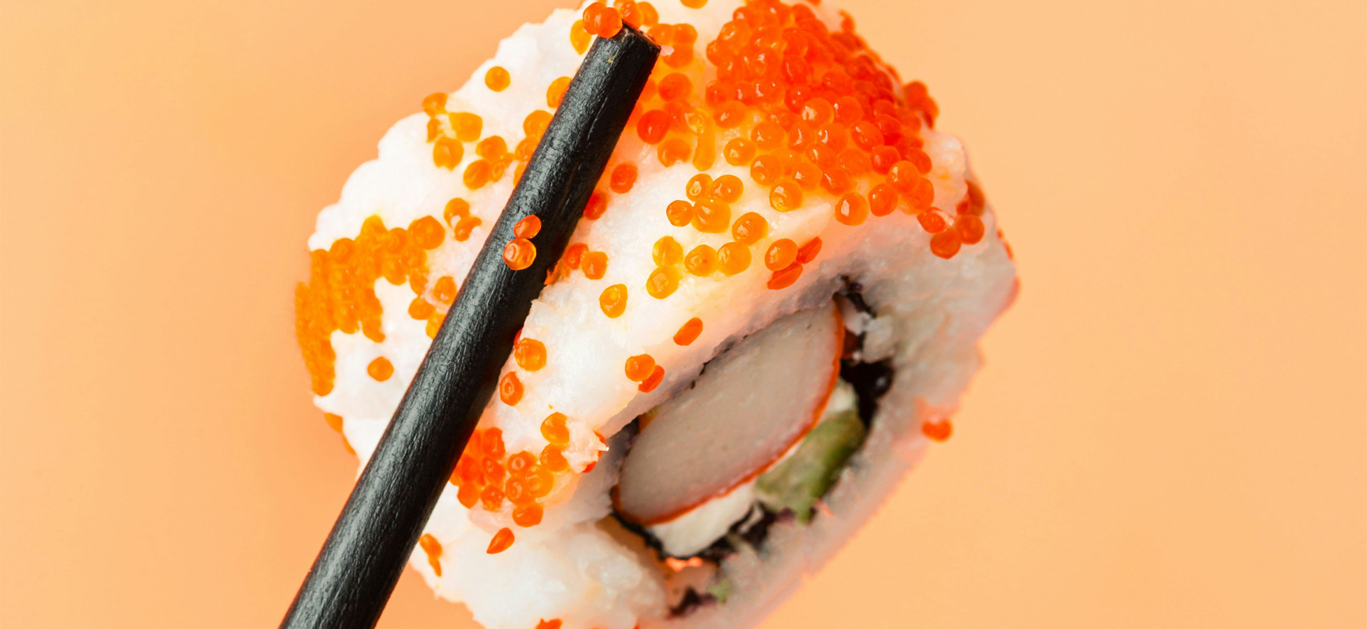 Ein Stück Sushi mit einer Füllung aus Fisch und Gemüse, umhüllt von Reis und orangefarbener Fischrogen. Es wird von schwarzen Essstäbchen gehalten und vor einem orangefarbenen Hintergrund platziert.