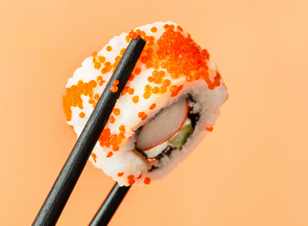Ein Stück Sushi mit einer Füllung aus Fisch und Gemüse, umhüllt von Reis und orangefarbener Fischrogen. Es wird von schwarzen Essstäbchen gehalten und vor einem orangefarbenen Hintergrund platziert.