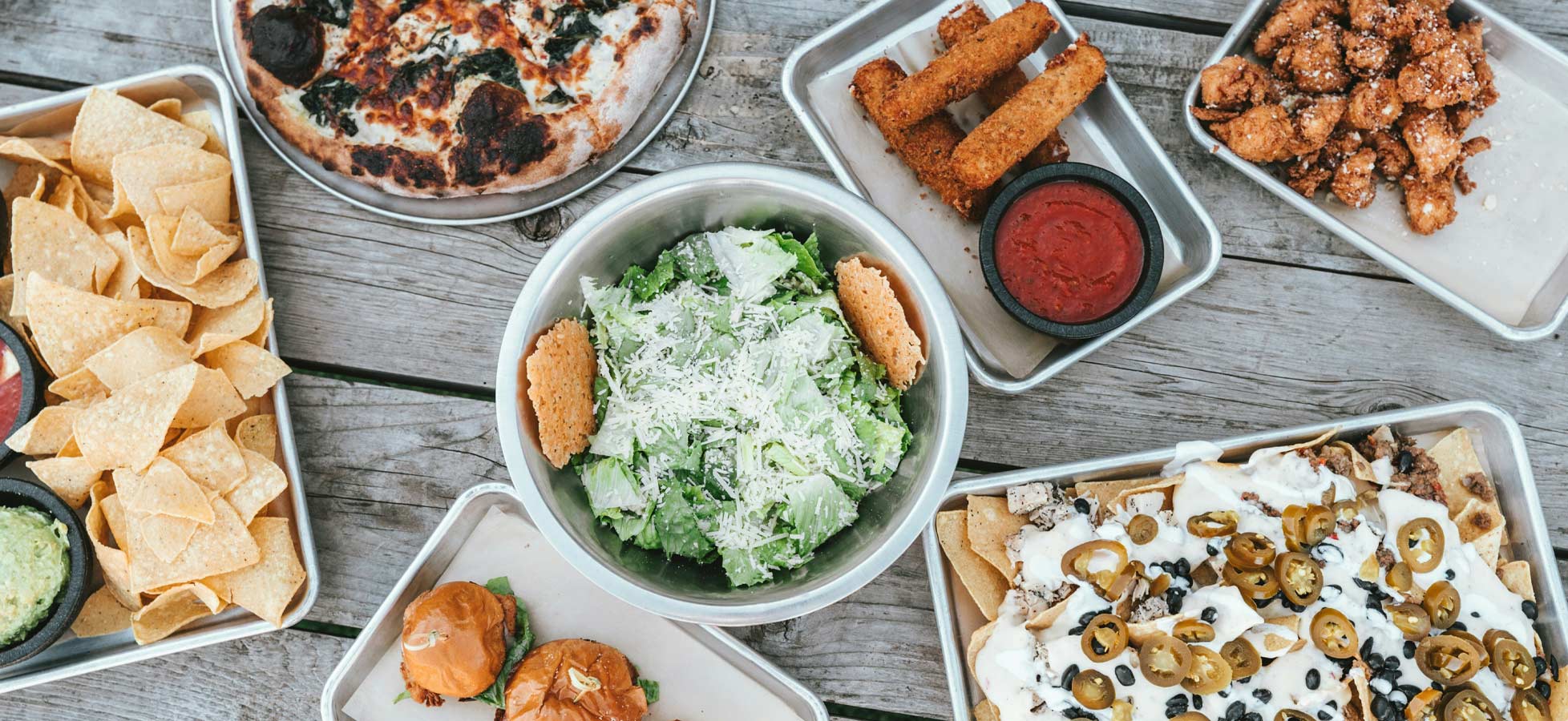 Eine Mischung aus verschiedenen Gerichten auf einem Tisch: knusprige Nachos mit Guacamole und Salsa, ein frischer Salat mit Parmesan, belegte Mini-Burger, eine Pizza und frittierte Snacks. Die Speisen sind ansprechend angerichtet und bieten eine bunte Auswahl für ein geselliges Essen.