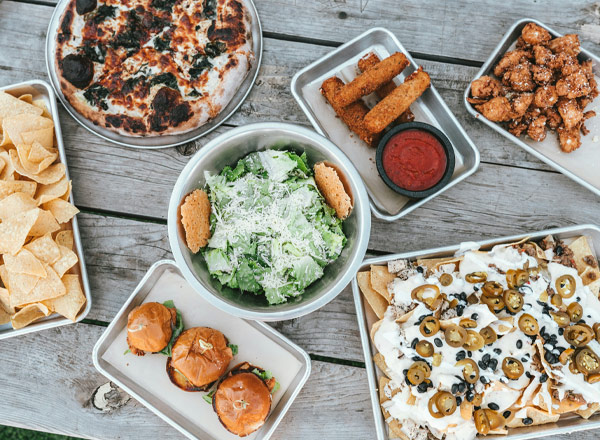 Eine Auswahl an Speisen auf einem Holztisch: eine Pizza, eine Schüssel mit Caesar-Salat, Nachos mit Jalapeños, Burger, Pommes und gebratene Hähnchenstücke mit einer Sauce. Die Gerichte sind in verschiedenen silbernen Behältern angerichtet.