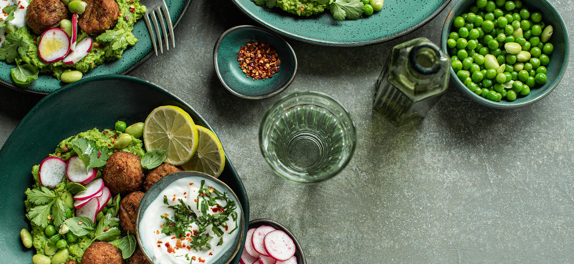 Ein gedeckter Tisch mit verschiedenen Schalen: Falaffel, Joghurt mit Kräutern, frische Radieschen und Avocado-Dip auf grünen Tellern. Zusätzlich sind ein Glas Wasser und eine Flasche Olivenöl zu sehen. Das Gesamtbild vermittelt eine gesunde, ansprechende Mahlzeit.
