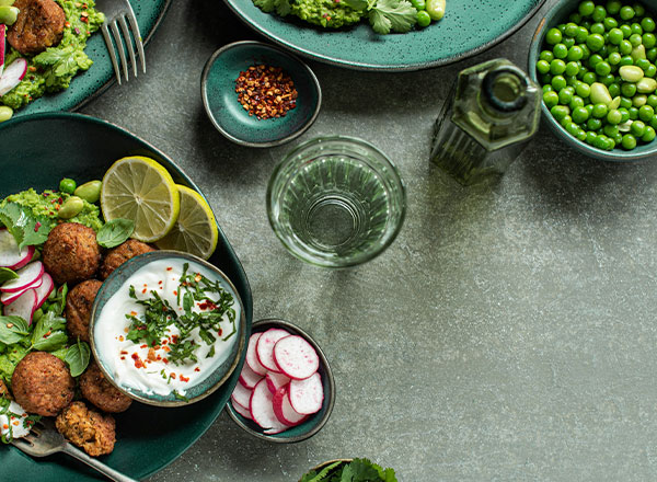 Ein gedeckter Tisch mit verschiedenen Schalen: Falaffel, Joghurt mit Kräutern, frische Radieschen und Avocado-Dip auf grünen Tellern. Zusätzlich sind ein Glas Wasser und eine Flasche Olivenöl zu sehen. Das Gesamtbild vermittelt eine gesunde, ansprechende Mahlzeit.