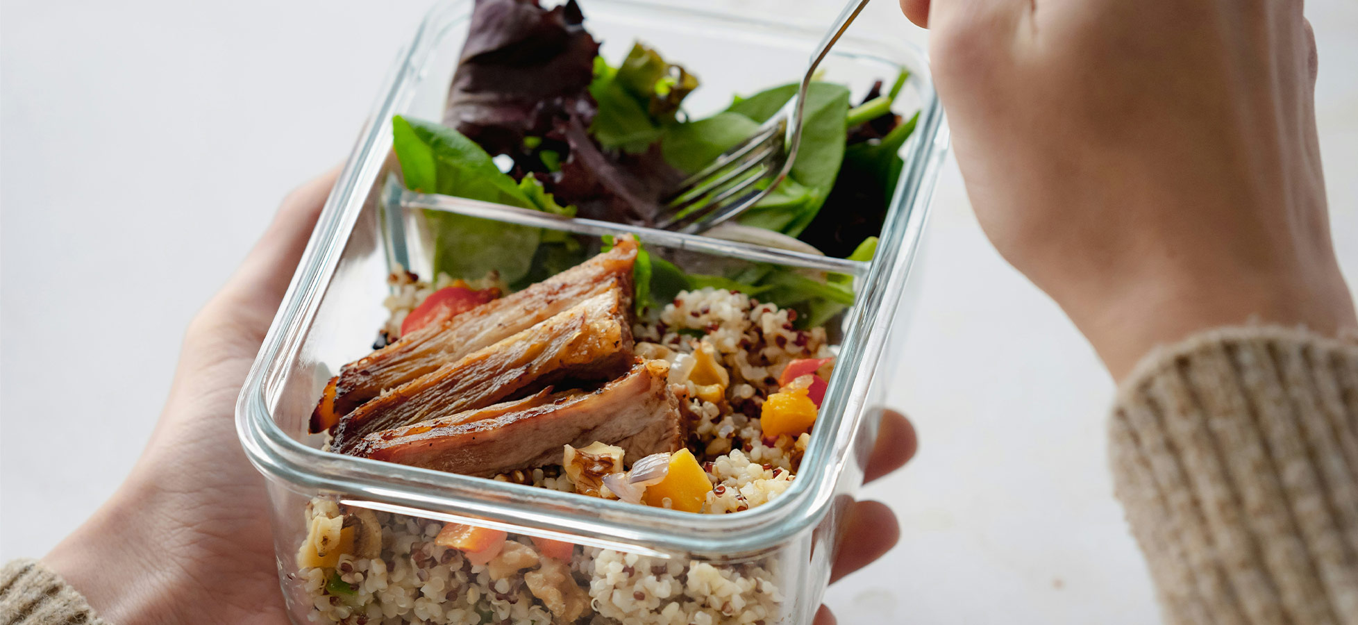 Eine Person hält eine durchsichtige Glasbox mit einem Mittagessen. Darin befinden sich Quinoa, bunte Gemüsewürfel und ein Stück gebratenes Fleisch. Zusätzlich liegt frischer grüner Salat obenauf. Eine Gabel ist in der anderen Hand der Person.